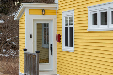 A bright red mailbox near the exterior door and porch of a yellow house with white trim.  There are a window and its reflection. Dark trees are at the rear of the house. The postal box is bright red.