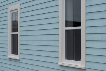The exterior wall of a baby blue country style house with narrow wood cape cod siding. There are two small double hung windows with white trim. Lace curtains are hanging in the closed glass windows.