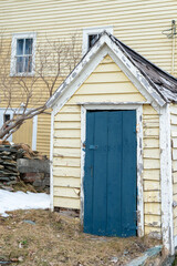 A small vintage pale yellow wooden clapboard shed with a vibrant blue closed shutter door. The trim on the building is painted white. There are rock walls and grass behind the building with snow.