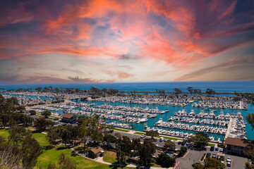 a stunning shot of the boats and yacht docked and sailing Dana Point Harbor with blue ocean water, lush green plants and trees on the hillside and powerful clouds at sunset in Dana Point California - obrazy, fototapety, plakaty