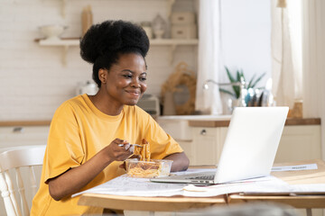 African girl eat and watch funny video, film on laptop, webinar for study or make conference call to friends or family. Busy female at break from remote work at home, studying distantly in kitchen