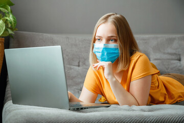 Woman in medical protective mask work using laptop at Home office lies on couch. Teen girl in mask Online learning education via laptop covid 19 lockdown time. Remote work in Coronavirus pandemic.