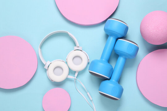 White stereo headphones and dumbbells on a blue background with pink circles. Minimalistic modern fitness layout