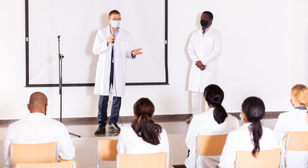 Fototapeta na wymiar Confident doctor wearing protective face mask to prevent viral infections standing with microphone on conference room stage, speaking to medical workers..