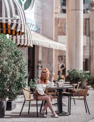 Stylish woman tourist in summer clothes with shorts and top blouse sits in street cafe in megapolis, city downtown, going to have a breakfast. Luxury and comfortable tourism season in UAE cityscapes