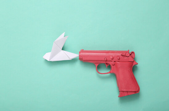 Pink pistol gun shooting origami dove on mint blue background. No war. Creative minimalistic layout. Flat lay. Top view