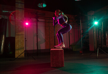 Fitness woman jumping on wooden box in pink green neon gradient light at modern gym. Sport,...