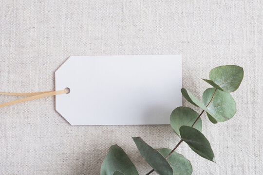 Blank tag card with eucalyptus leaves on pastel natural linen fabric. Mockup for design, branding, sale or invitation. Top view, flat lay, copy space