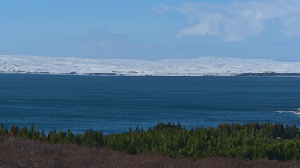 Beautiful view of blue colored lake Þingvallavatn, largest natural lake of Iceland, in Þingvellir national park with rugged, snow-covered mountains and forest in front in winter season on sunny day.