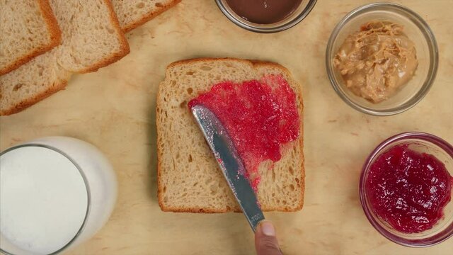 Spreading strawberry jam with a butter knife on a slice of tasty brown bread - popular breakfast. Various healthy tasty breakfast items like a glass of milk  peanut butter  fruit jam  and slices of...