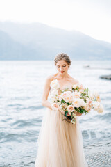 Fototapeta na wymiar Pensive bride with a bouquet of flowers stands on the shore of Lake Como against a background of mountains
