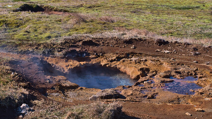 Small hot spring with steam in geothermal area Geysir, Haukadalur, Iceland, part of Golden Circle, with orange deposits and green colored moss on sunny winter day.