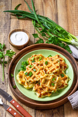 Not sweet waffles with potatoes, peas, bell peppers, cheese are served with sour cream and herbs on a plate on a rustic table.