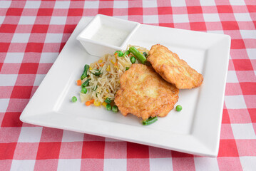 Fried fish with vegetable salad served in a white square plate over red plaid table cloth.