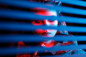 Portrait of a young woman in neon light, looking through the blinds. A trendy portrait with colored lighting and striped shadows.