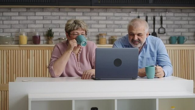 cheerful elderly married pair is drinking tea at home kitchen and viewing movie or pictures on laptop display