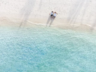 Poster Im Rahmen Young couple traveler sitting and relaxing at beautiful tropical white sand beach with wave foam and transparent sea, Summer vacation and Travel background Top view from drone © Kittiphan