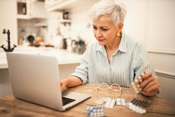 Indoor shot of serious attractive retired female sitting at table with portable computer keyboarding, holding blisters pack, looking for information about painkillers, suffering from headache
