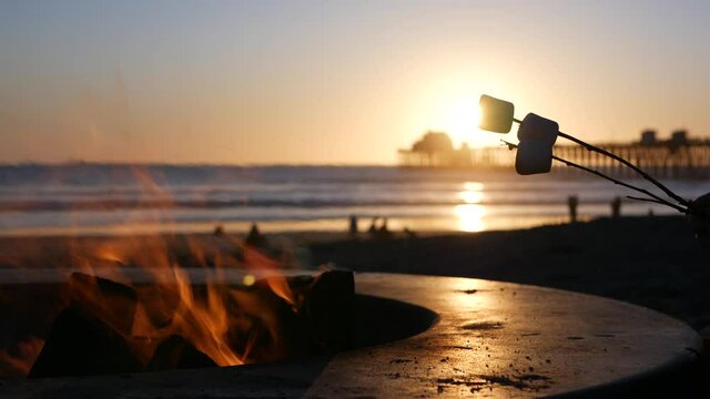 Campfire pit by Oceanside pier, California USA. Camp fire on ocean beach, bonfire flame in cement ring place for bbq, sea water waves. Heating, roast or toast marshmallow on stick. Romantic sunset sky