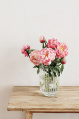 Floral still life scene. Pink peonies flowers, bouquet in glass vase on wooden table. White wall. Selective focus, blurred background. Wedding, birthday celeberation concept. Lifestyle vertical photo