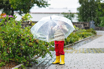 Little blond kid boy walking with big umbrella outdoors on rainy day. Preschool child having fun...