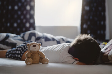 Cute Teddy bear sitting on bed next to child boy with morning light.Peaceful scene of New day Young Kid lying in bed with happy brown bear and the curtain opening with natural light shining through.