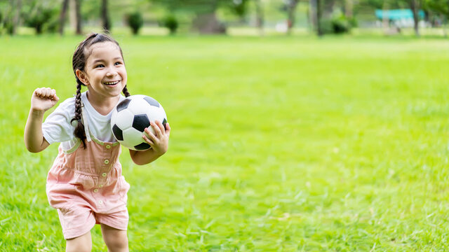 Smiling little girl standing holding the soccer ball at green football field in summer day. Portrait of little girl athlete playing with a ball at stadium. Active childhood concept. Copy space