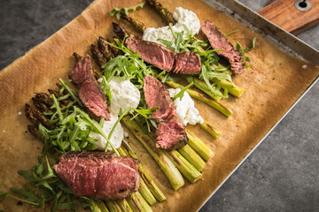 Gerösteter grüner Spargel mit medium rare Rinder Steak Streifen, Rucola, Büffel Mozzarella und Olivenöl auf Backblech mit Backpapier, Holzbrett, Besteck und rustikal dunkel Stein Hintergrund
