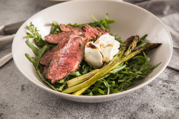Gerösteter grüner Spargel mit medium rare Rinder Steak Streifen, Rucola, Büffel Mozzarella, Olivenöl und Balsamico in Schale mit Leinen Serviette, Besteck und rustikal Stein Hintergrund
