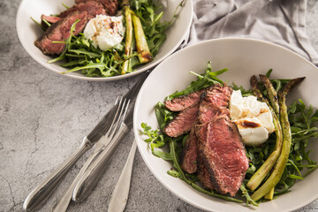 Gerösteter grüner Spargel mit medium rare Rinder Steak Streifen, Rucola, Büffel Mozzarella, Olivenöl und Balsamico in Schale mit Leinen Serviette, Besteck und rustikal Stein Hintergrund
