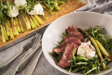 Gerösteter grüner Spargel mit medium rare Rinder Steak Streifen, Rucola, Büffel Mozzarella und Balsamico in Schale und auf Backblech mit Backpapier, Besteck und rustikal Stein Hintergrund