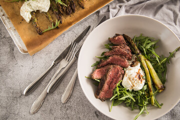 Gerösteter grüner Spargel mit medium rare Rinder Steak Streifen, Rucola, Büffel Mozzarella und Balsamico in Schale und auf Backblech mit Backpapier, Besteck und rustikal Stein Hintergrund