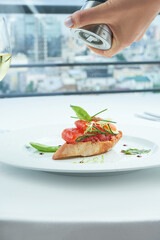 Delicious Italian bruschetta with salmon, cucumber on a white baguette, served in a white plate on a white tablecloth. A woman's hand is grinding pepper on a bruschetta.