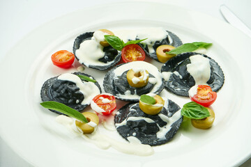 Fototapeta na wymiar Delicious cuttlefish ink ravioli stuffed with shrimps and scallops, served with white sauce, cherry tomatoes and olives in a white plate on a white tablecloth. Black ravioli pasta