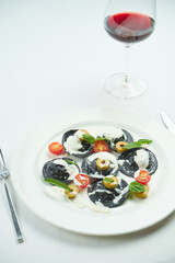 Delicious cuttlefish ink ravioli stuffed with shrimps and scallops, served with white sauce, cherry tomatoes and olives in a white plate on a white tablecloth. Black ravioli pasta