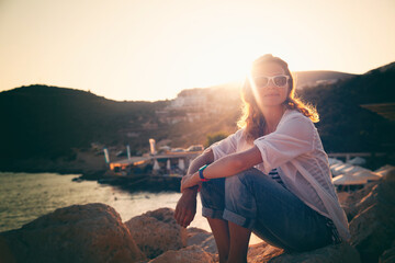 Beautiful young woman ib white shirt and sunslasses sitting by the sea at sunset in the sun