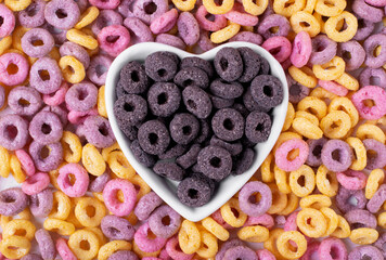 Chocolate cereal loops in heart shaped bowl and multicolored rings scattered around. Breakfast food