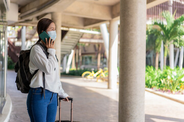 Fototapeta premium travel, woman in white long-sleeved shirt and jeans, wearing a mask. using mobile phone during travel with suitcase in the left hand and backpack