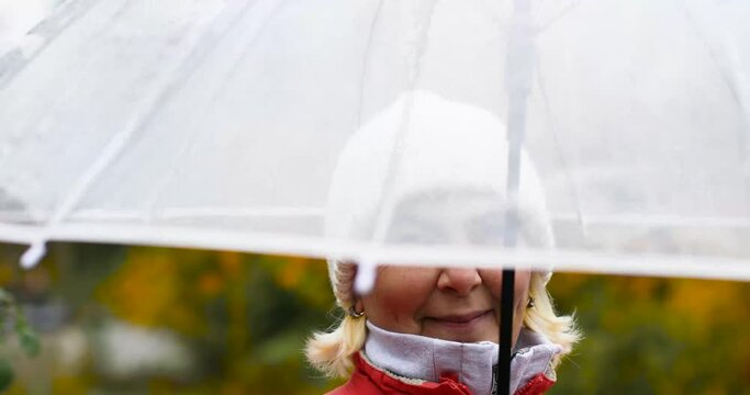 Adult woman 50 years old in a red jacket and under a transparent umbrella enjoying the rainy weather. Autumn mood, yellow leaves