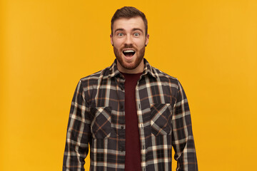 Fototapeta premium Surprised handsome young man in checkered shirt with beard and opened mouth looks amazed and shouting over yellow background