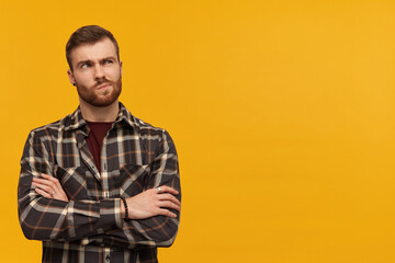Thoughtful sceptical young man in checkered shirt with beard keeps arms crossed and thinking over yellow background Looking away to the side - Powered by Adobe