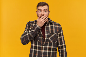 Obraz premium Scared shocked young man in plaid shirt with beard covered his mouth by hand and looks stunned over yellow background