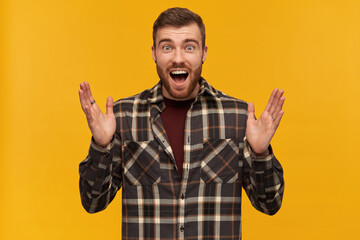 Naklejka premium Happy amazed young man in checkered shirt with beard and opemed mouth keeps hands raised and looks surprised over yellow background