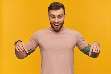 Happy handsome young man in pink tshirt with beard and tattoo on hand looking and pointing down by two fingers over yellow background
