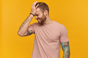 Disappointed embarrassed young bearded man with tattoo and eyes closed in pink tshirt keeps hand on...