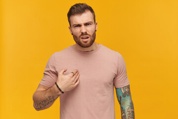 Irritated handsome young man in pink tshirt with beard and tattoo on hand points at himself by hand and looks annoyed over yellow background