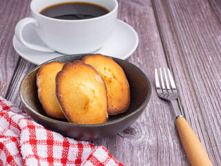 Madeleine homemade traditional French small cookies on black a dish and a cup of coffee placed on a wooden background. Lemon glaze madeleines. Seashell sweet cakes.