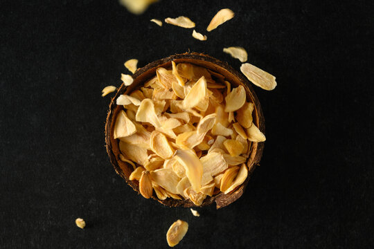 Dry Flakes Of Garlic In A Coconut Bowl, Flakes Falling From Above On A Dark Gray Background, Top View