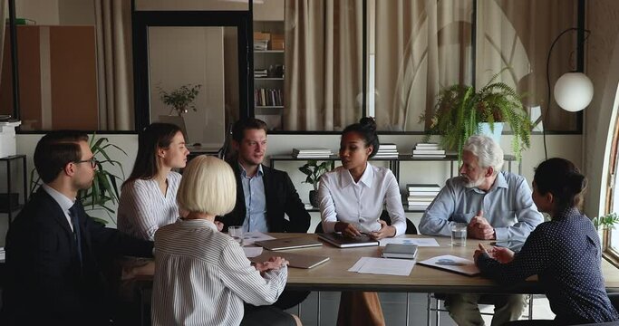 Confident young african american business lady sharing opinion about problem solution or brainstorming project ideas with motivated diverse older and young colleagues at briefing meeting in office.