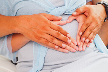 Beautiful and warm moment of father and mother putting their heart shaped hands together on growing baby bump expecting their cute baby at home Love and care and family lifestyle concept